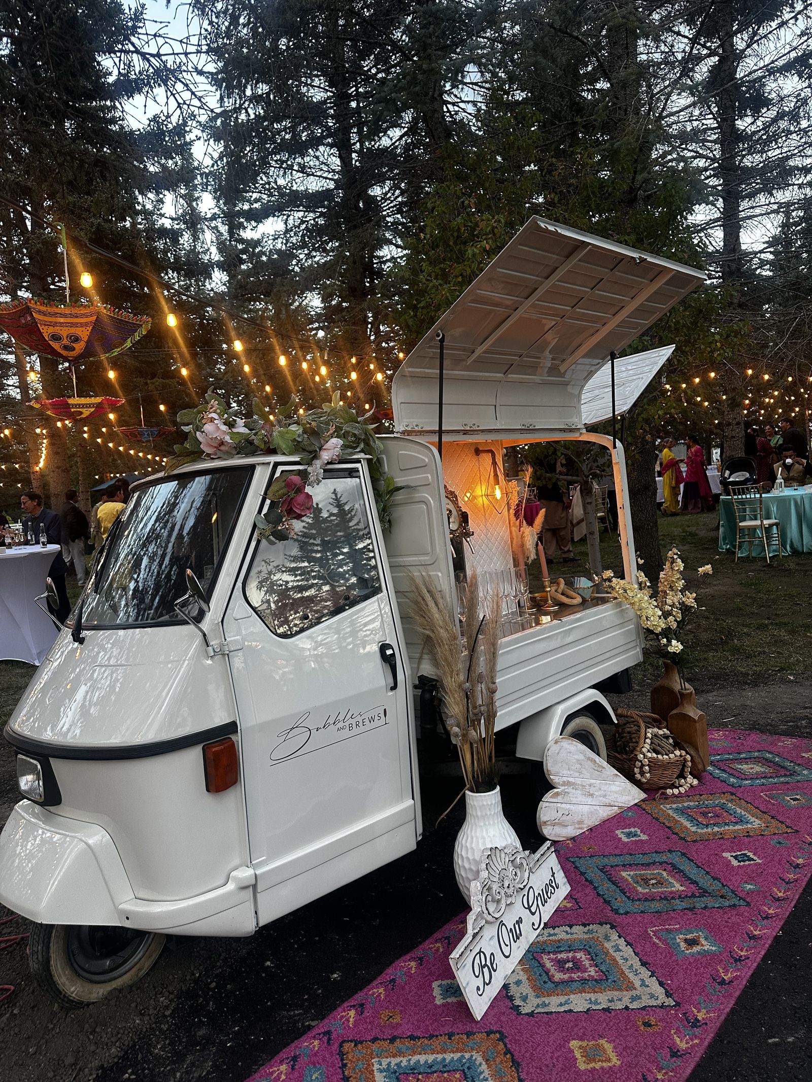 Bubbles and Brews mobile bar at an evening reception under string lights with a 'Be Our Guest' sign