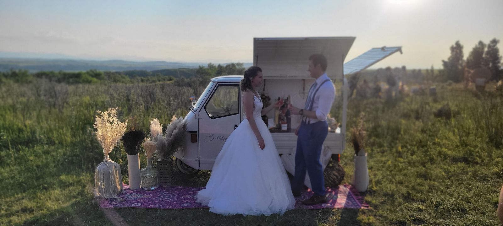 Wide panoramic view of a wedding couple beside the mobile bar at golden hour in the Alberta foothills