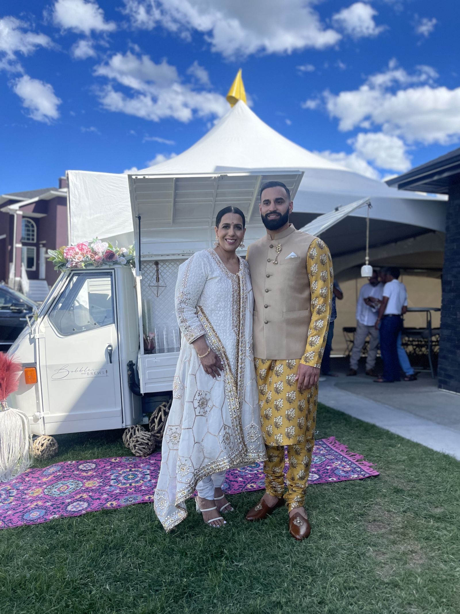 Couple celebrating in traditional formal wear in front of the mobile bar under an event tent